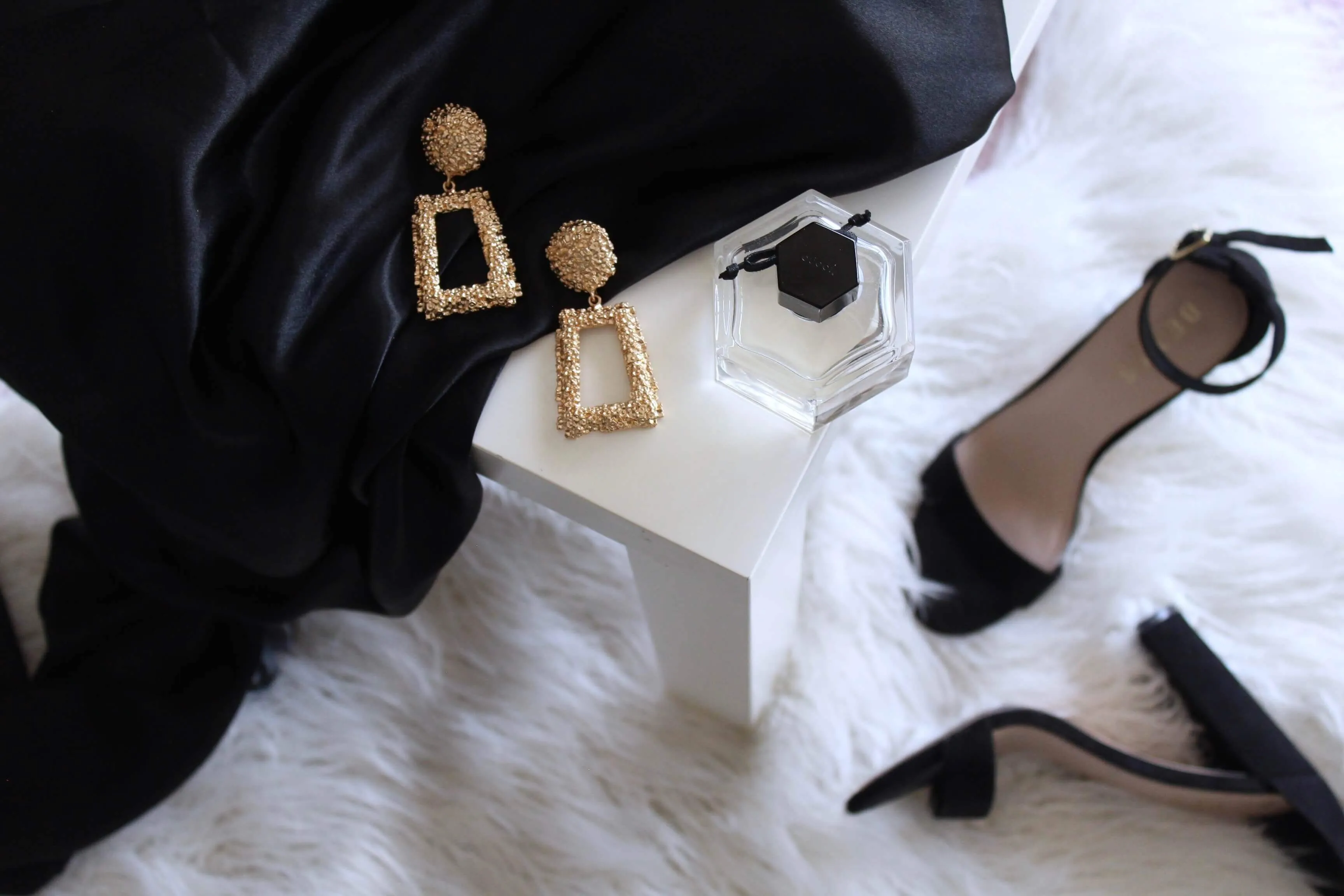 Gold-tone earrings, a bottle of perfume, a sandal and black velvet garmet draped across a luxurious white shag rug.